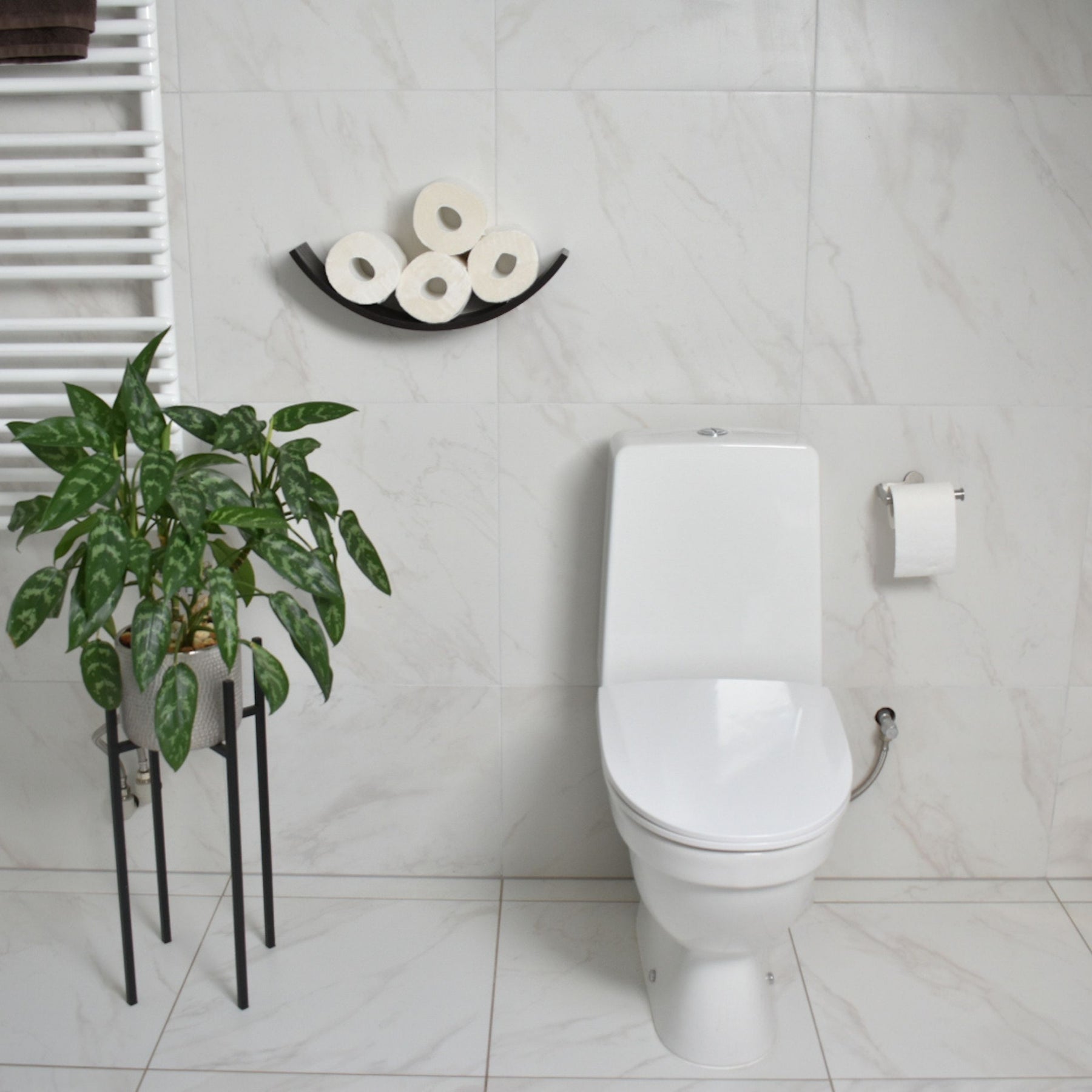 Front view of dark wood curved toilet paper holder mounted on a bathroom wall, displaying four rolls of toilet paper. The bathroom features minimalist design with a plant, modern fixtures, and a clean, marble tile backdrop.