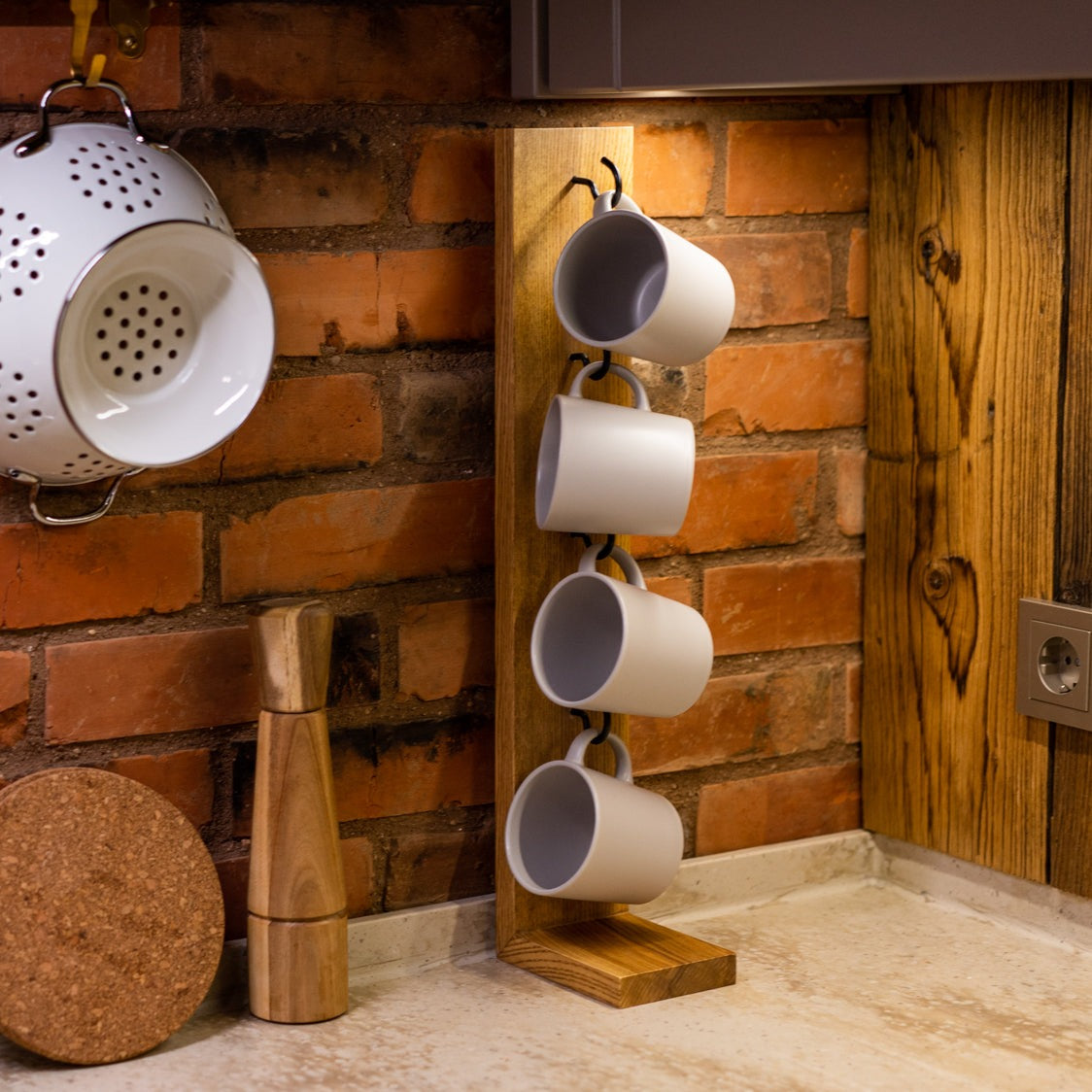Wooden mug holder with hooks, displaying four white mugs, set on a countertop with a brick backsplash and wooden accents, creating a cozy kitchen atmosphere.