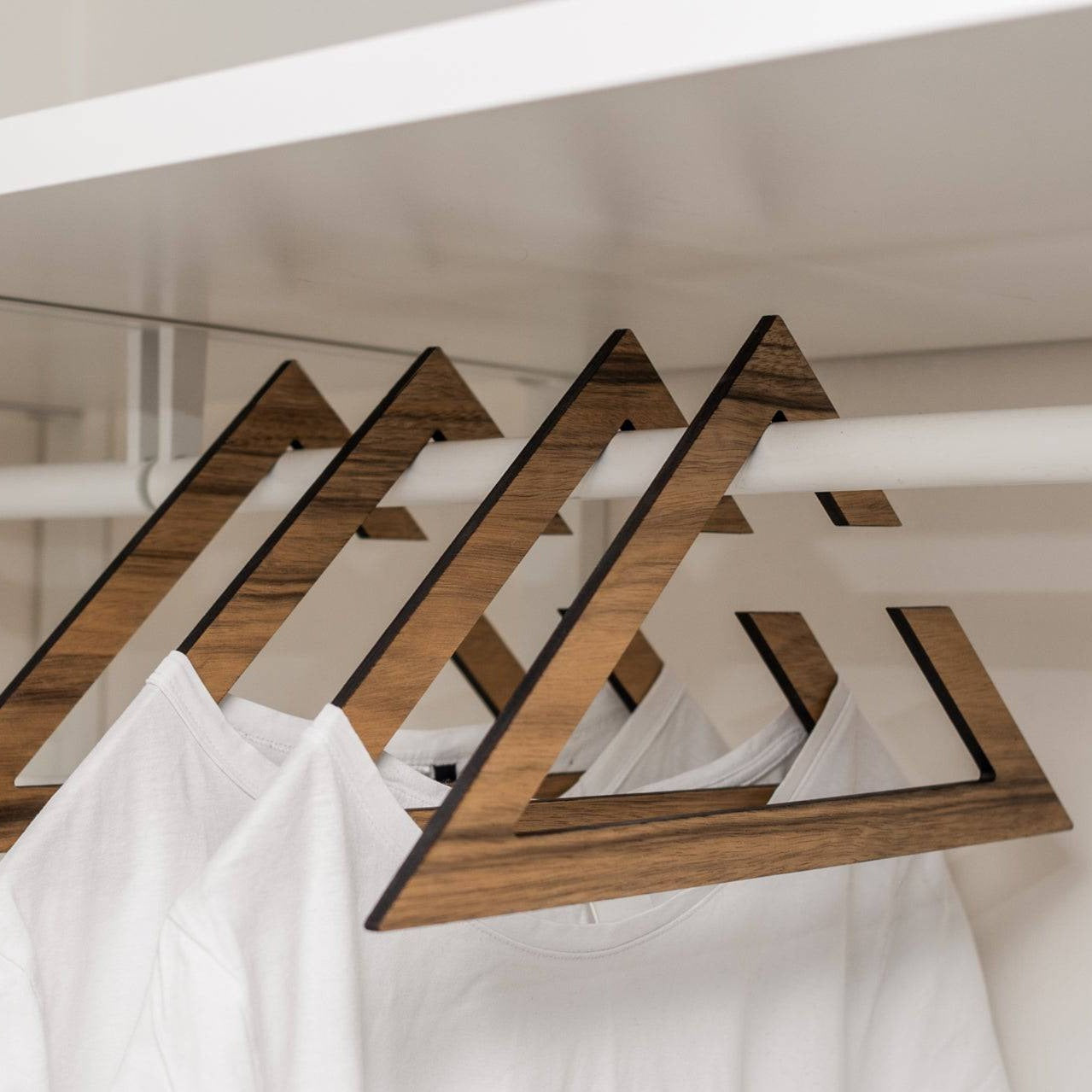 Stylish wooden hangers with a geometric triangular design, showcasing a rich paldau finish. The hangers hold white t-shirts in a minimalist closet setup.