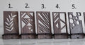 Wooden phone stands in walnut finish with unique geometric cutout designs: 1. Leaf pattern, 2. Abstract triangles, 3. Diagonal lines, 4. Geometric shapes, 5. Circular cutouts. Each stand holds a phone at an angle on a smooth surface.