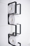 Close-up of a wall-mounted black toilet paper holder with a geometric design, displaying three rolls of toilet paper stacked vertically.
