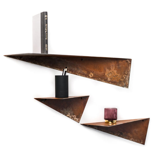 Three triangular metal shelves with a rustic, oxidized finish. The top shelf holds a book, the middle shelf holds a black cylindrical object, and the bottom shelf holds a pink glass candle holder and a small decorative item.