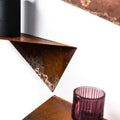 Close-up of a triangular metal shelf with a rustic, oxidized finish and textured surface, holding a black cylindrical object and a pink glass candle holder, set against a white wall.