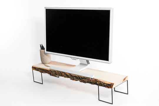 Contemporary computer screen elevated on a natural edge wooden platform, featuring a textured bark detail and supported by thin black metal supports. A minimalist ceramic cup with pens sits on the left.
