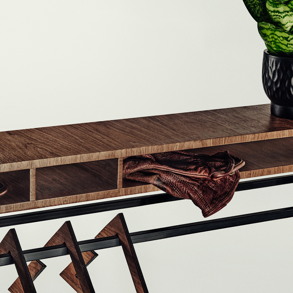 Close-up of a dark wood console table with open cubbies, a draped brown leather accessory, geometric wooden legs, and a potted plant on a light neutral background.