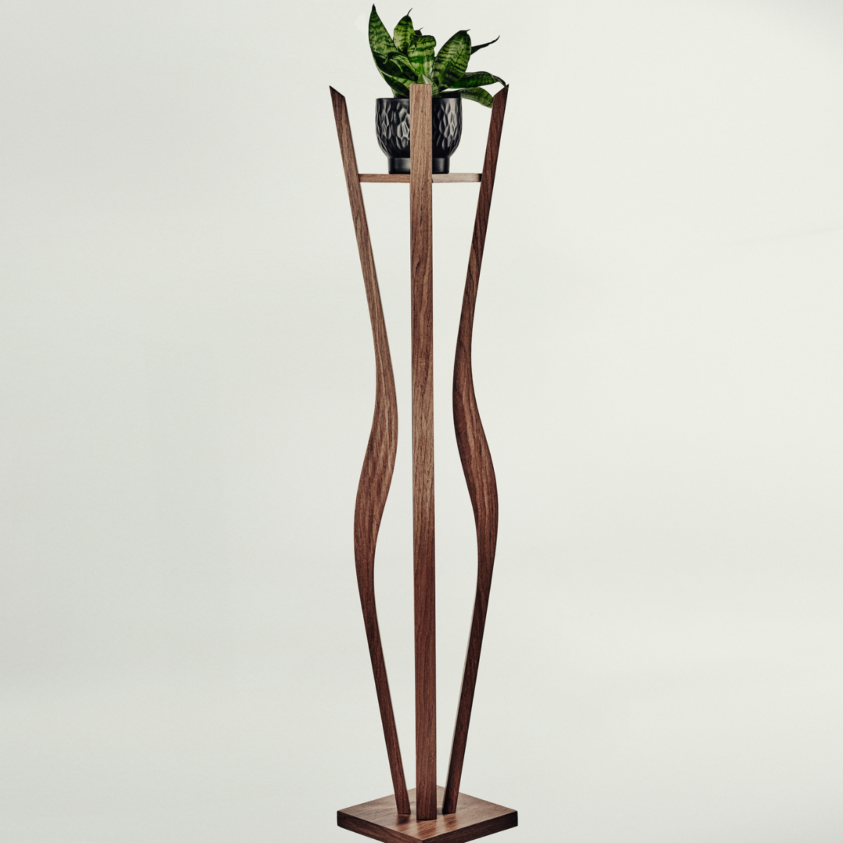 A tall sculptural wooden plant stand with a rectangular base and two curved vertical supports, holding a small potted green plant on a narrow shelf. The stand appears to be made of medium-to-dark brown wood, photographed against a plain light gray studio background.