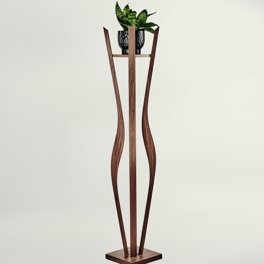 A tall sculptural wooden plant stand with a rectangular base and two curved vertical supports, holding a small potted green plant on a narrow shelf. The stand appears to be made of medium-to-dark brown wood, photographed against a plain light gray studio background.