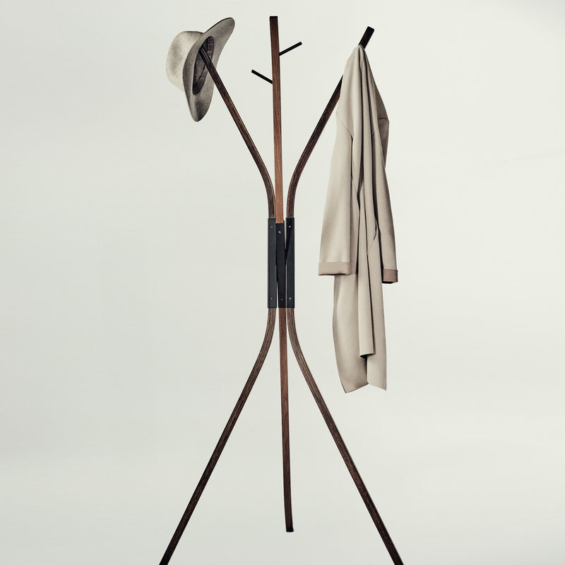 A modern wooden coat rack with three legs and curved top arms, shown against a plain light gray background. A hat is hanging on the left hook and a light beige coat is hanging on the right.