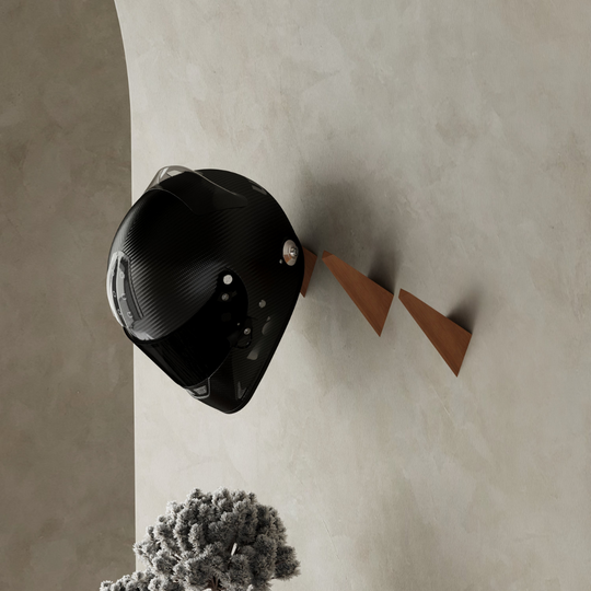Wall-mounted wooden hooks with a triangular design, used to hold a black motorcycle helmet, set against a neutral-toned wall with decorative foliage below.