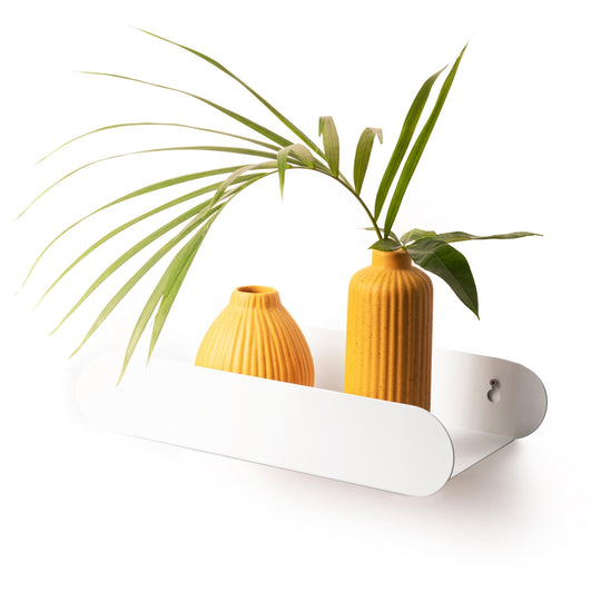 White, capsule-shaped metal shelf with a glossy finish, mounted on a wall with screws. It holds two orange, ribbed vases with green palm leaves, highlighting a minimalist design.