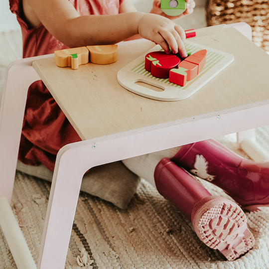 Child in a terracotta dress and pink wellies arranges wooden toy fruits and vegetables on a pale wood and soft pink play table.