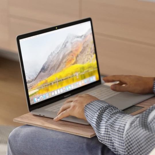 A person using a laptop on a wooden sofa tray. Features: Made in Europe, 10 color options, natural wood, non-slip fabric, folds on one side. Ewart Woods logo is visible in the corner.