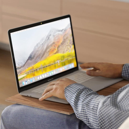 A person using a laptop on a wooden sofa tray. Features: Made in Europe, 10 color options, natural wood, non-slip fabric, folds on one side. Ewart Woods logo is visible in the corner.