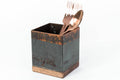 Square utensil holder with a rustic copper finish and wooden base, containing copper-colored forks and a spoon. The holder is placed against a plain white background.