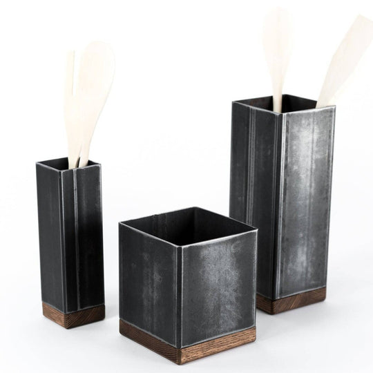 Set of three black metal rectangular utensil holders with wooden bases in varying heights, each holding light-colored wooden utensils, against a plain white background.