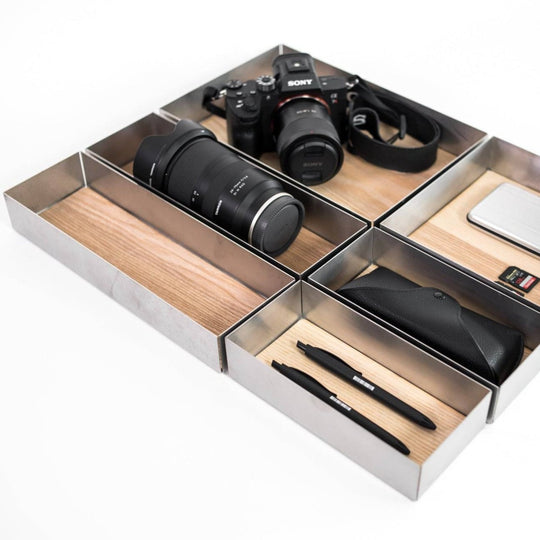 A modular set of storage boxes with wooden bases and metal sides. The compartments are used to organize various items like a camera, lens, pens, and a sunglasses case, showcasing their versatile storage potential.