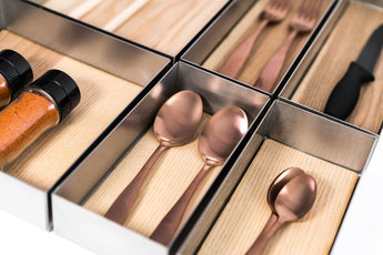 Organized metal trays containing various kitchen utensils, including two copper spoons, forks, a knife, and spice containers filled with red seasoning. The utensils are neatly arranged on a wooden surface.