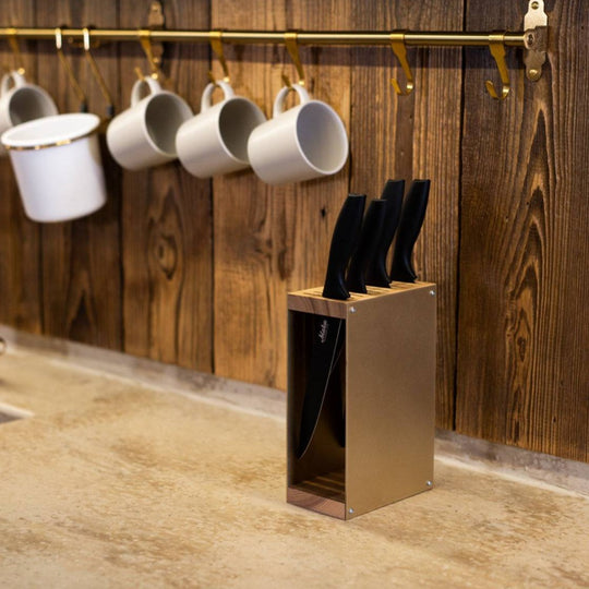 Gold-colored knife block with a wooden top and base, holding black-handled knives. The knife block is placed on a kitchen countertop with a rustic wooden wall in the background.