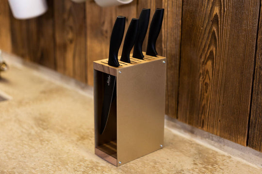 Gold-colored knife block with a wooden top and base, holding black-handled knives. The knife block is placed on a kitchen countertop with a rustic wooden wall in the background.