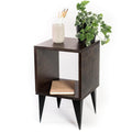 Dark-stained wooden side table with a square open compartment, black tapered legs, and a minimalist design. Decorated with a plant, a ceramic container holding paintbrushes, and a cork notebook placed inside