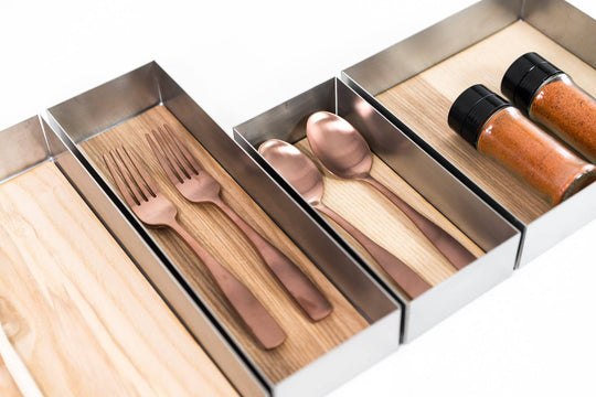 Four metal compartments with wooden bases holding rose gold forks, spoons, and two spice jars filled with red seasoning, arranged in a sleek kitchen organizer.