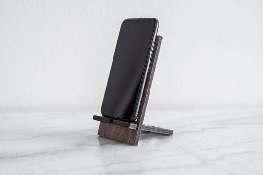 Walnut wooden phone stand with a simple, modern design, securely holding a phone at an angle on a smooth surface.