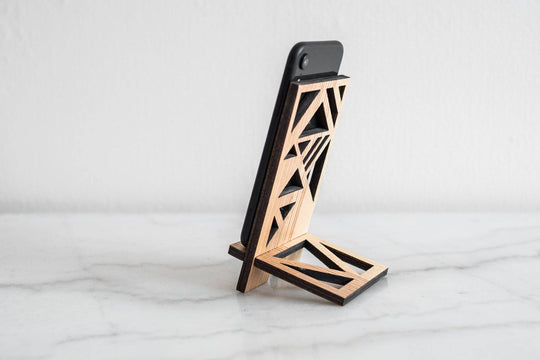 Wooden phone stand in a combination of light and dark wood finishes with a geometric design featuring intricate triangular patterns, holding a phone at an angle on a smooth surface.