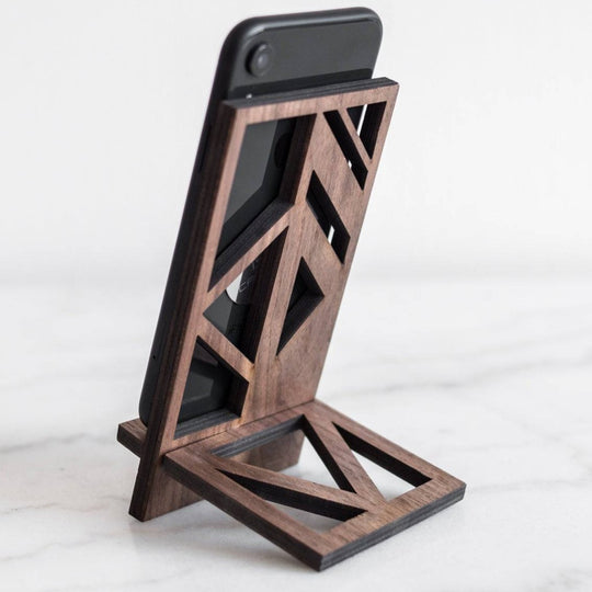 Wooden phone stand in walnut finish with a geometric design, holding a phone at an angle, placed on a smooth surface.