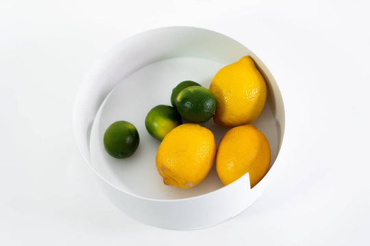 Minimalist white metal bowl with a geometric design, filled with fresh lemons and limes, placed on a clean white background.