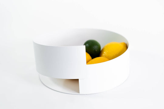 Contemporary white metal bowl with a layered, modern design, holding vibrant lemons and limes on a white background.
