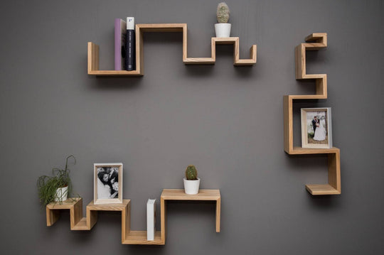 Three tiered wooden shelves with natural grain, asymmetrically mounted on a gray wall, displaying books, plants, and pictures.
