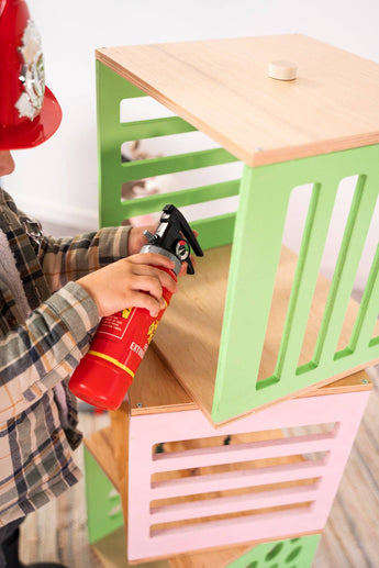 Child playing with toy fire extinguisher next to stacked wooden blocks in green and pink, featuring grid-like cutouts and flat wooden tops.
