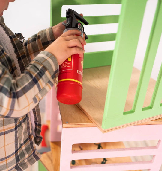 Child holding a toy fire extinguisher, pointing it at a green wooden block with cut-out slats, stacked on top of pink and green blocks with grid designs.