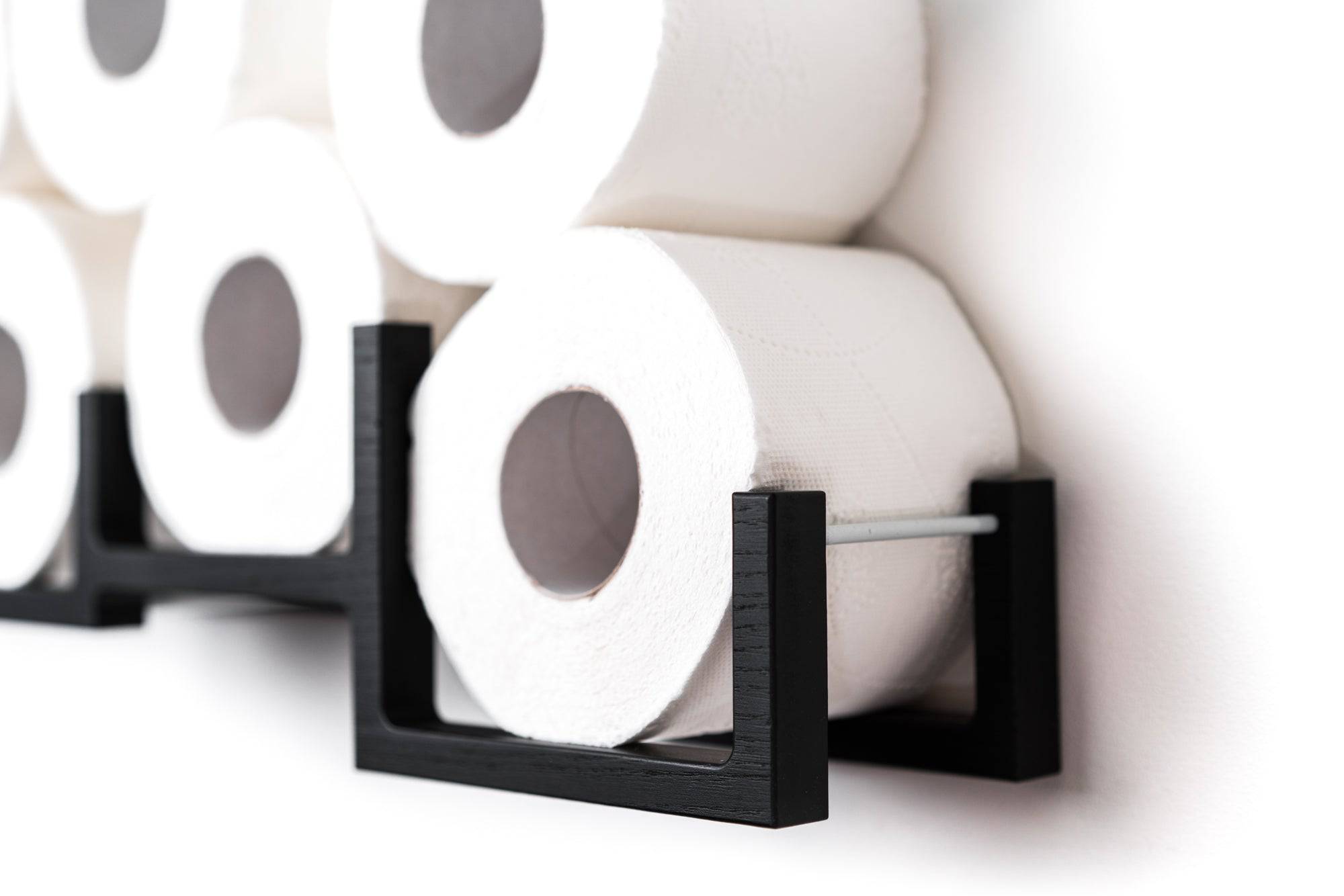 Close-up of a black wooden wall-mounted toilet paper holder, displaying several rolls of toilet paper. The holder has a modern, geometric design with square frames and a metal bar for support.