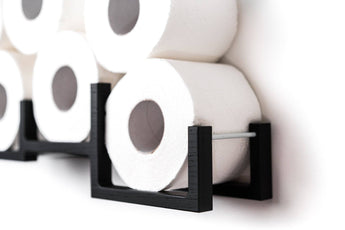 Close-up of a black wooden wall-mounted toilet paper holder, displaying several rolls of toilet paper. The holder has a modern, geometric design with square frames and a metal bar for support.