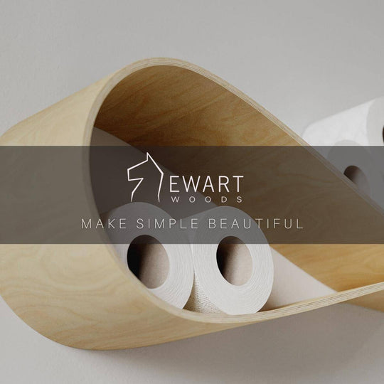 Close-up of a floating birch wood drop-shaped wall shelf showing rich wood texture, holding toilet paper rolls. The image includes the EWART WOODS logo and the tagline Make Simple Beautiful in the center.